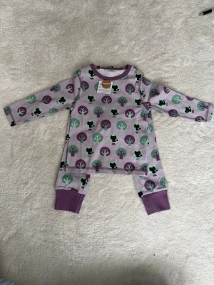 Lila bodyset med träd och kattmotiv - Sött tvådelat set med långärmad body och byxor i lila toner. Setet har ett lekfullt mönster med träd i grönt och lila samt små svarta katter. Muddar i lila vid ärm- och benslut. Tillverkat i mjuk ekologisk bomull, perfekt för mysiga dagar.