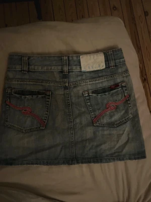 Vintage  jeanskjol - Säljer en blå jeanskjol i kort modell med två bakfickor som har rosa broderade detaljer. Kjolen har bälteshällor och klassisk femficksdesign. Köpte på secondhand i Polen 