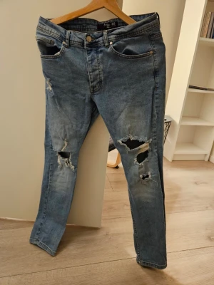 Blå jeans - Säljer ett par blå slim fit jeans från H&M med flera slitningar och hål på båda benen. Jeansen har klassisk femficksmodell och normal midja. Materialet är denim i bomull.