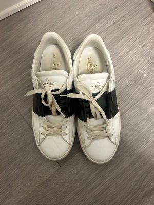 Valentino vita sneakers med svart detalj - Snygga vita sneakers från Valentino Garavani med bred svart läderdetalj över snörningen. Klassisk låg modell med rund tå och vita skosnören. Perfekta för dig som gillar stilrena och exklusiva sneakers.