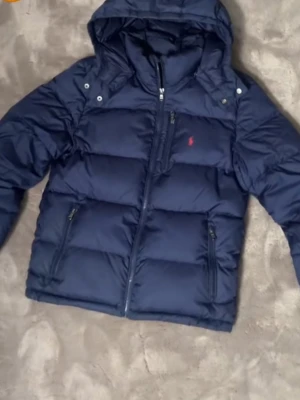pufferjacka Ralph Lauren priset kan diskuteras  - Snygg mörkblå pufferjacka från Ralph Lauren med huva och dragkedja framtill. Jackan har två sidofickor med dragkedja och en bröstficka. Ikoniska röda Polo-loggan på bröstet. Perfekt för kalla dagar och riktigt skön att bära.