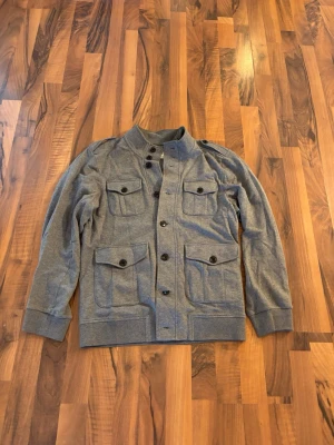 Grå utilityjacka/tröja  med fickor - Snygg grå utilityjacka med fyra stora fickor framtill och svarta knappar. Jackan har ståkrage och ribbade muddar vid ärmsluten. Tillverkad i mjukt tyg, perfekt för lager på lager och streetwear-stil. Perfekt att ha över en tröja men funkar också över en schysst tisha! Kan passa S också beroende på vad man har för kläder under. 