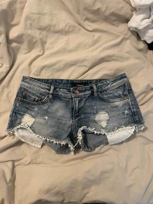 jeansshorts  - Snygga blå jeansshorts med slitna detaljer och vit spetskant längst ner. Shortsen har klassisk femficksdesign, knappgylf och råa kanter för en trendig look. Perfekta för sommaren och ger en avslappnad vibe.