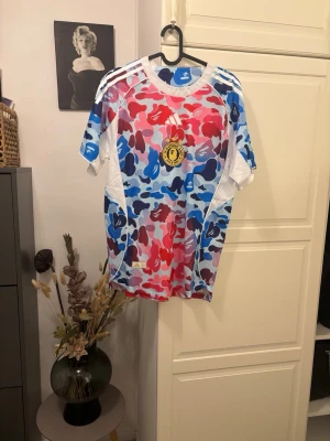 Adidas x Bape camo fotbol jersey  - Säljer en färgstark Adidas x Bape t-shirt med camomönster i rött, blått och vitt. Tröjan har klassiska Adidas-ränder på axlarna och meshpaneler på sidorna för extra ventilation. Perfekt för dig som gillar streetwear och unika plagg. 