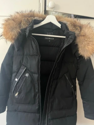 Svart dunjacka  Cedrico - Snygg svart dunjacka från Cedrico med stor huva och lyxig pälsdetalj runt kanten. Jackan har två snedställda fickor med dragkedja och tryckknapp, samt justerbar midja och huva. Perfekt för kalla vinterdagar och riktigt varm.