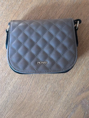 Grå quiltad handväska från Picard - Snygg grå handväska från Picard i quiltat konstläder med guldfärgad logga framtill. Väskan har rundade kanter, lockstängning och en justerbar axelrem. Perfekt storlek för det viktigaste och en stilren look.