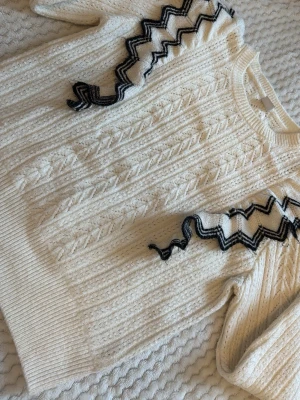 Vit stickad tröja med zigzag-detaljer - Säljer en krämvit stickad tröja med strukturmönster och unika svarta zigzag-detaljer på axlarna. Tröjan har rund halsringning och långa ärmar. Perfekt för dig som gillar att sticka ut med coola detaljer och mjuka material. En liten fläck som inte syns direkt men villl visa att det finns en litet fläck vid handen!🤍
