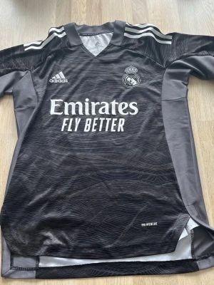 Real Madrid målvaktströja Adidas svart - Snygg svart Real Madrid målvaktströja från Adidas med Courtois och nummer 1 på ryggen. Tröjan har vita detaljer, klubbmärke på bröstet och Emirates Fly Better-tryck. Tillverkad i Primeblue-material med gråa paneler på sidorna och axlar.