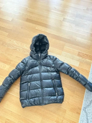 Svart pufferjacka från Colmar - Säljer en svart pufferjacka från Colmar med huva och två dragkedjeförsedda fickor. Jackan har en glansig finish och klassisk quiltad design. Colmar-logga på ärmen. Perfekt för kalla vinterdagar och riktigt snygg till streetwear-stilen.