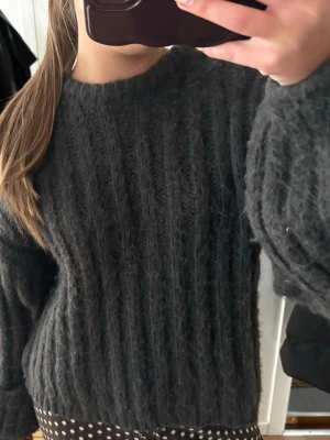 Mörkgrå fluffig stickad tröja - Mysig mörkgrå stickad tröja med fluffig känsla och breda ribbade ränder. Tröjan har rund halsringning och långa, lite pösiga ärmar. Perfekt för kalla dagar och enkel att styla till olika outfits.