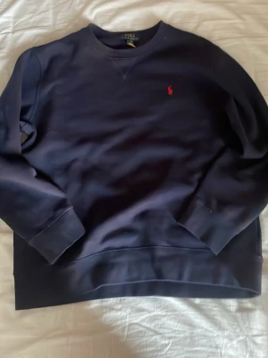Mörkblå sweatshirt från Polo Ralph Lauren - Mörkblå sweatshirt från Polo Ralph Lauren med klassisk rund halsringning och ribbade muddar. Tröjan har det ikoniska röda Polo-logotypen broderad på bröstet. Perfekt oversized look och mjukt material för en chill vibe.