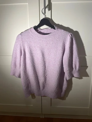 Ljuslila stickad tröja med puffärm - Supersöt ljuslila stickad tröja med puffiga korta ärmar och ribbad rund hals. Tröjan har en mjuk och fluffig struktur som känns riktigt mysig. Perfekt för dig som vill ha en trendig och bekväm look till vardags.