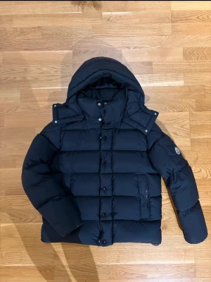 Moncler vezere "skannar" - Snygg svart pufferjacka från Moncler med stor huva och två dragkedjefickor framtill. Jackan har en patch med Moncler-logga på ena ärmen och quiltad design. Perfekt för kalla dagar och riktigt trendig look.