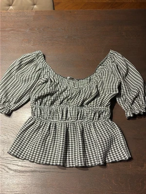 Rutig blus med puffärm - Söt svartvit rutig blus med bred resår i midjan och puffiga trekvartsärmar. Blusen har en lätt v-ringad halsringning och volang nedtill för en trendig look. Perfekt för dig som gillar en feminin och avslappnad stil. XS/S