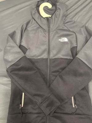 Svart Grå Hoodie North Face - Snygg hoodie från The North Face i svart och mörkgrått med dragkedja framtill och två fickor med dragkedja. Loggan är tryckt i vitt på bröstet och ryggen. Hoodien har en klassisk huva och är tillverkad i ett mjukt, stretchigt material.