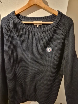 Mörkblå stickad tröja från Morris - Säljer en mörkblå stickad tröja från Morris med rund halsringning och diskret logga på bröstet. Tröjan har ett grovt stickat mönster och långa ärmar, perfekt för dig som gillar klassisk och stilren design.