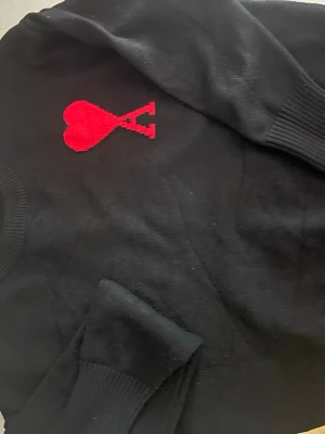 Svart stickad tröja från Ami Paris - Säljer en svart stickad tröja från Ami Paris med deras ikoniska röda hjärta och A broderat på bröstet. Tröjan har rund halsringning och ribbade muddar vid ärmslut och nederkant. Perfekt för dig som gillar stilrena plagg med en twist.