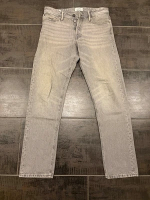 Grå raka jeans från Jack & Jones - Snygga grå jeans från Jack & Jones med rak passform och klassisk femficksdesign. Jeansen har knappgylf och är tillverkade i ett slitstarkt denimtyg. Perfekta för en avslappnad och trendig look. Storlek 31/34