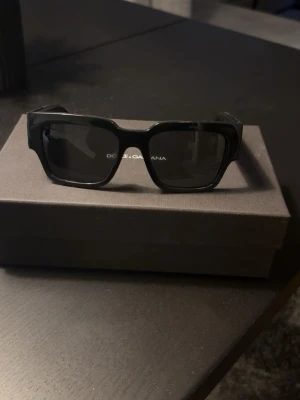 Svarta solglasögon Dolce & Gabbana - Snygga svarta solglasögon från Dolce & Gabbana med rektangulära bågar och breda skalmar där loggan syns tydligt i vitt. Glasen är mörka och bågarna är tillverkade i blank syntet. Endast använd några gånger, nypris 3000