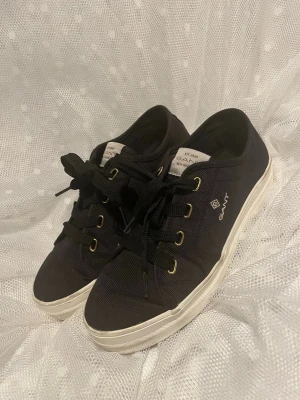 Svarta låga sneakers från Gant - Snygga svarta sneakers från Gant med vita sulor och guldfärgade öljetter. Klassisk låg modell med textilöverdel och svarta skosnören. Perfekta för en clean och stilren look. Stl. 40