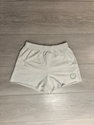 Vita shorts med SMILEY-tryck - Säljer ett par vita shorts med elastisk midja och grönt SMILEY-tryck baktill samt en liten smiley framtill. Sköna och avslappnade.
