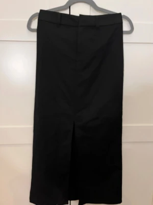 Svart långkjol med slits - Klassisk svart kostymkjol med hög midja och slits framtill. Kjolen har bälteshällor och rak passform, perfekt för en stilren look. Tillverkad i ett slätt material som ger en snygg siluett. 