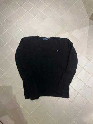 Svart kabelstickad tröja Polo Ralph Lauren - Kabelstickad svart tröja från Polo Ralph Lauren med rund halsringning och lila broderad logga på bröstet. Långärmad modell i mjukt material, perfekt för lager på lager. Klassisk och stilren design som funkar till det mesta.
