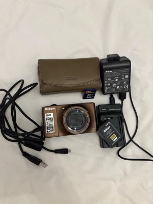 Nikon Coolpix brun kompaktkamera - Säljer en brun Nikon Coolpix digitalkamera med 10x optisk zoom, 14.2 megapixlar och 3-tums LCD-skärm. Kameran har HD-videoinspelning och medföljer laddare, USB-kabel, batteri, SD-kort och ett brunt Nikon-fodral. Perfekt för dig som vill ha en smidig kompaktkamera.