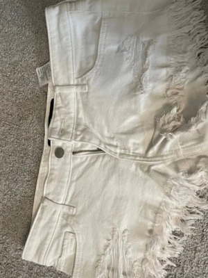 Hollister lågmidjade jeans shorts - Säljer ett par vita jeansshorts med hög midja och fransiga kanter. Shortsen har slitna detaljer framtill, klassiska fickor både fram och bak samt dragkedja och knappstängning. Perfekta för sommaren och riktigt snygga till en enkel topp.