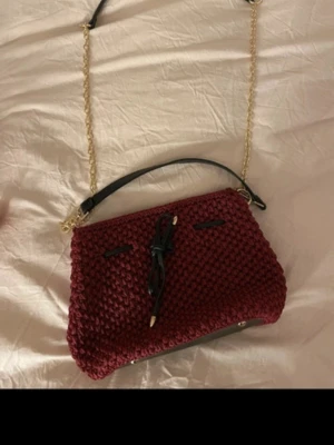 Vinröd stickad handväska med kedja - Snygg vinröd handväska i stickat material med svart handtag och guldfärgad kedjerem. Väskan har en dekorativ svart snörning framtill och detaljer i svart konstläder. Perfekt för dig som vill ha en unik accessoar. egengjord väska