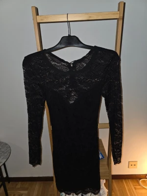 Svart spetsklänning med öppen rygg - Snygg svart kort klänning i spets med lång ärm och öppen rygg. Klänningen har figurnära passform och fina spetsdetaljer både fram och bak. Perfekt för dig som vill ha en elegant och trendig look.