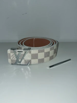 L.V bälte - Snyggt bälte från L.V med klassiskt rutigt mönster i beige och vitt. Bältet har en stor silverfärgad L.V som spänne och är gjort i skinn. Perfekt accessoar för att lyfta vilken outfit som helst.