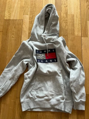Grå hoodie från Tommy Jeans XS - Snygg grå hoodie från Tommy Jeans med stor logga framtill i blått, vitt och rött. Klassisk känguruficka och justerbar huva med dragsko. Tillverkad i mjuk bomullsblandning som är perfekt för chill dagar. Ribbade muddar vid ärmslut och nederkant.