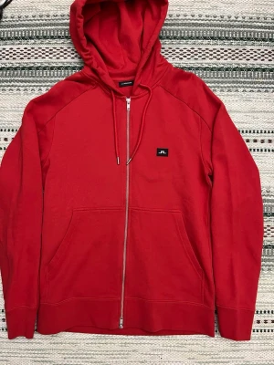 Röd hoodie med dragkedja från J.Lindeberg - Snygg röd hoodie från J.Lindeberg med dragkedja framtill, huva med dragsko och känguruficka. Liten svart patch med vit logga på bröstet. Tillverkad i mjuk bomullsblandning, perfekt för en avslappnad stil.