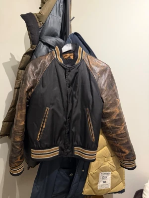 Svart PRADA bomberjacka med bruna läderärmar - Snygg svart bomberjacka med slitna bruna läderärmar och matchande detaljer på krage, muddar och midja. Jackan har två fickor framtill med bruna läderkanter och stängs med knappar. Perfekt för dig som gillar retrostil och streetwear.