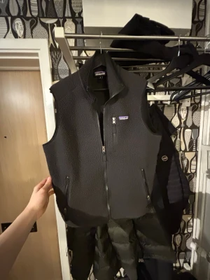 Svart fleeceväst från Patagonia -  svart fleeceväst från Patagonia med dragkedja framtill och två praktiska fickor med dragkedja. Klassisk modell utan ärmar och med hög krage. Perfekt lager-på-lager-plagg för kyliga dagar. Priset är ej hugget i sten!