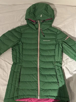 Grön dunjacka Peak Performance - Snygg grön dunjacka från Peak Performance med huva och dragkedja framtill. Jackan har rosa detaljer på insidan och runt snörningen samt en randig dragkedja. Perfekt för kalla vinterdagar och har en modern, sportig look.