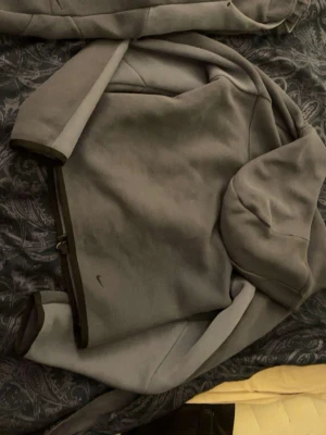 Grå hoodie från Nike med dragkedja - Säljer en grå hoodie från Nike med dragkedja framtill och huva. Hoodien har diskret Nike-logga på bröstet och svarta detaljer vid ärmslut och dragkedja. Perfekt för en avslappnad och sportig stil.