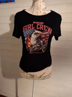 Svart Girl Crew t-shirt med tryck - Cool svart t-shirt med färgstarkt tryck av en örn, motorcykel och amerikansk flagga samt texten 'WEEKEND GIRL CREW'. T-shirten har rund hals, korta ärmar och en något croppad passform. Perfekt för dig som gillar statement-plagg.