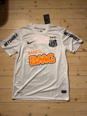 Vit Santos Neymar Jr matchtröja Nike - Santos FC matchtröja i vitt med Neymar Jr och nummer 11 på ryggen. Tröjan har tryckta sponsorer, klubbmärke och Nike-logga. Tillverkad i lätt och ventilerande material, perfekt för fotboll eller streetwear.