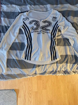 Vit långärmad zip-up topp med tryck. - Säljer en vit långärmad topp med svarta detaljer och tryck framtill. Toppen har tryck, samt svarta ränder längs sidorna. Materialet känns mjukt och stretchigt. Köpte den på Camden Market i London, därför vet ej vilket märke. Perfekt för er som gillar 2000’s stil. Blixtlåset går att få ner men blir sedan lite trögt på botten. Den funkar men behöver dra lite hårdare. 