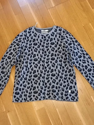 Grå leopardmönstrad stickad tröja - Snygg grå stickad tröja från Three Sixty Five med svart leopardmönster. Tröjan har rund halsringning och långa ärmar. Perfekt för dig som vill ha en trendig och bekväm look med lite edge. Materialet känns mjukt och passar bra till jeans.