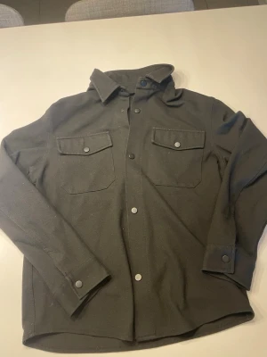 Svartovershirt från Zara - Stilren svart overshirt från Zara med två bröstfickor och tryckknappar framtill. Skjortjackan har klassisk krage och raka ärmar med knapp vid manschetten. Perfekt lager-på-lager-plagg i mjukt tyg.
