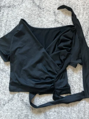 Svart omlottopp med knytband - En svart omlottopp med korta ärmar och långa knytband i midjan. Toppen har en djup v-ringning och är tillverkad i ett mjukt, stretchigt material som passar perfekt till träning eller dans. Original pris 150kr