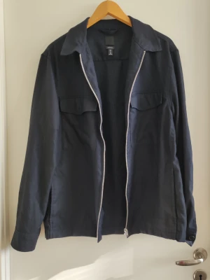 Marinblå overshirt med dragkedja - Stilren marinblå overshirt med silverfärgad dragkedja framtill och två bröstfickor med lock. Klassisk krage och raka ärmar med knapp vid ärmslut. Perfekt lager-på-lager-plagg i ett mjukt bomullsliknande material.