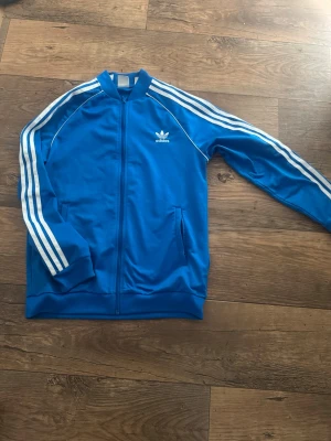 Blå Adidas track jacket med vita ränder - Snygg blå track jacket från Adidas med klassiska vita ränder längs ärmarna och Adidas-logga på bröstet. Dragkedja framtill, ribbade muddar och två fickor. Perfekt för en sportig och retro look.