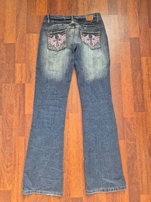 Blå bootcut jeans med broderi - Säljer ett par blå bootcut jeans med rosa broderade detaljer på bakfickorna. Jeansen har låg midja och kontrastsömmar i rosa. Materialet är klassisk denim.