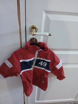 Röd vindjacka med nummer 49 - Röd vindjacka med vita och marinblå detaljer samt tryckt nummer 49 på bröstet. Jackan har dragkedja framtill, reflexdetaljer på ärmarna och huva. Perfekt för blåsiga dagar.