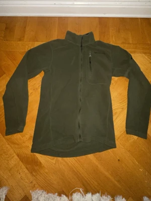 Mörkgrön peak performance kofta - Mörkgrön fleecejacka från Haglöfs med hel dragkedja och hög krage. Jackan har en bröstficka med dragkedja och långa ärmar. Perfekt för lager-på-lager och outdoor-aktiviteter. Materialet är mjukt och värmande fleece.