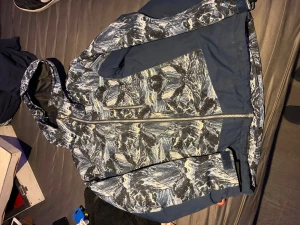 Mönstrad vindjacka från North Bend - Snygg vindjacka från North Bend med blått och vitt abstrakt mönster, hel dragkedja och huva. Jackan har två sidofickor med dragkedja och justerbara muddar vid ärmsluten. Knappt använd.  Storlek 158/164.